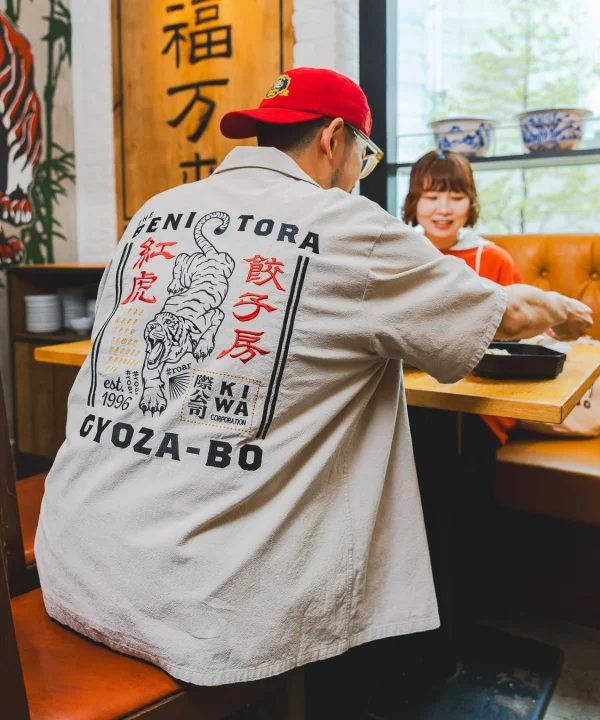 中華料理店で食事中のカップルの後ろ姿。男性は虎と「紅虎餃子房」の文字が入ったシャツを着ており、女性は笑顔で彼を見ている。テーブルには料理が置かれ、和やかな雰囲気が漂う。