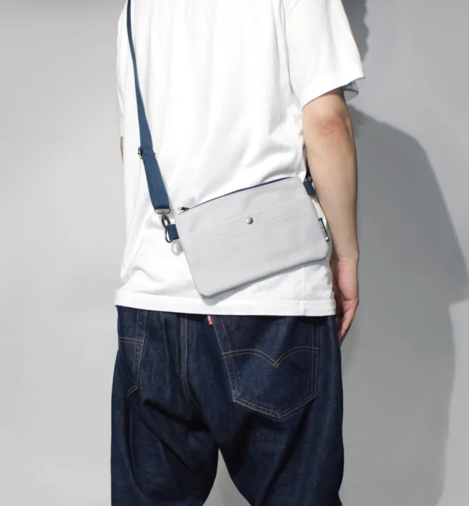 白いTシャツと濃い色のジーンズを着用し、ライトグレーのサコッシュを斜めがけにした男性の後ろ姿です