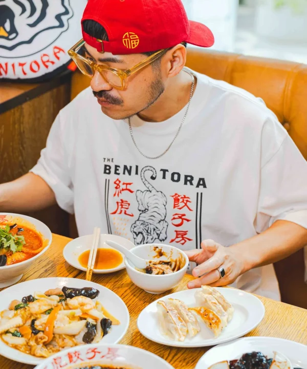 一人の男性が中華料理店で食事を楽しんでいます。テーブルには餃子や麺料理、炒め物などが並び、男性は特徴的な眼鏡と赤いキャップを着用しています。