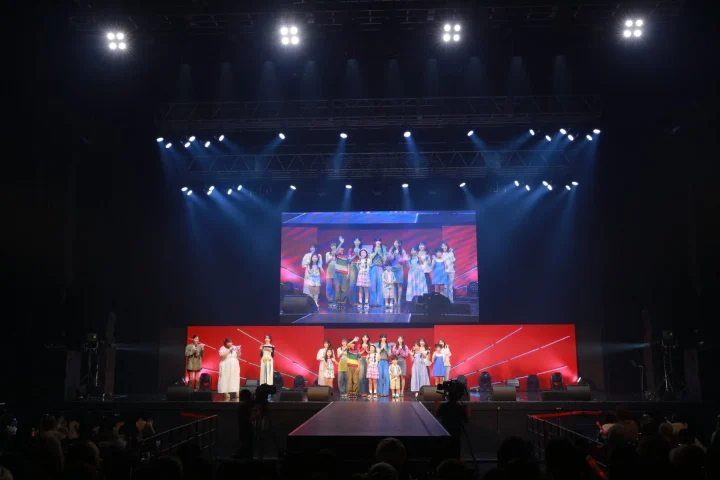複数の出演者（大人と子供）がステージに立ち、観客の前でパフォーマンスを行っているライブイベントの様子。