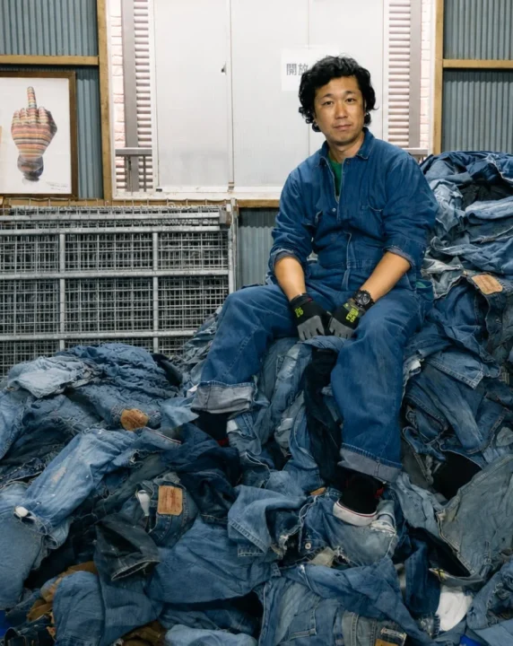 デニムのつなぎを着た男性が、大量に積み上げられたジーンズの山の上に座っている