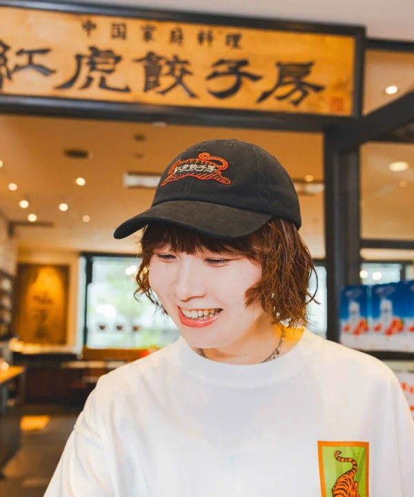 中華料理店「紅虎餃子房」の看板を背景に、黒いキャップと白いTシャツを着たショートヘアの女性が笑顔を見せています。キャップには店名がデザインされ、Tシャツにも虎のイラストが見えます。