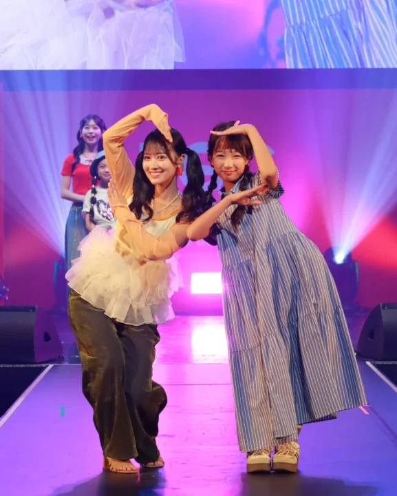 明るいステージ上で、2人の女性が笑顔でハートのポーズを取っています。