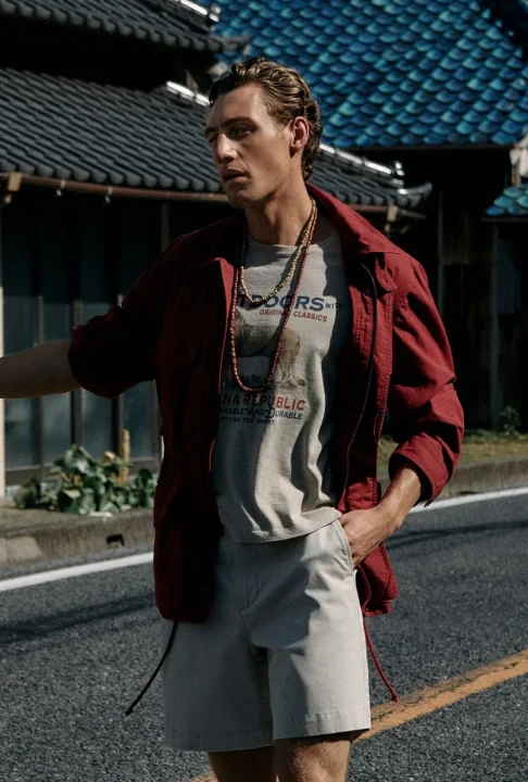 伝統的な日本家屋を背景に、路上に立つ男性モデル。赤いジャケットとグレーのTシャツ、ベージュのショートパンツを着用し、ネックレスを重ね付けした夏のカジュアルファッションを披露している。