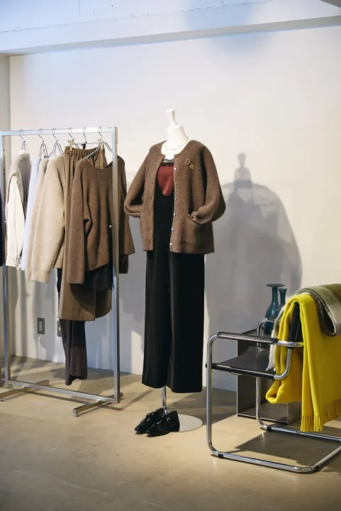 マネキンがブラウンのカーディガンと黒いパンツを着用し、隣にはニュートラルカラーの洋服が並んだラックがあります。モダンな椅子には黄色いブランケットがかけられ、アパレル店舗のディスプレイのようです。
