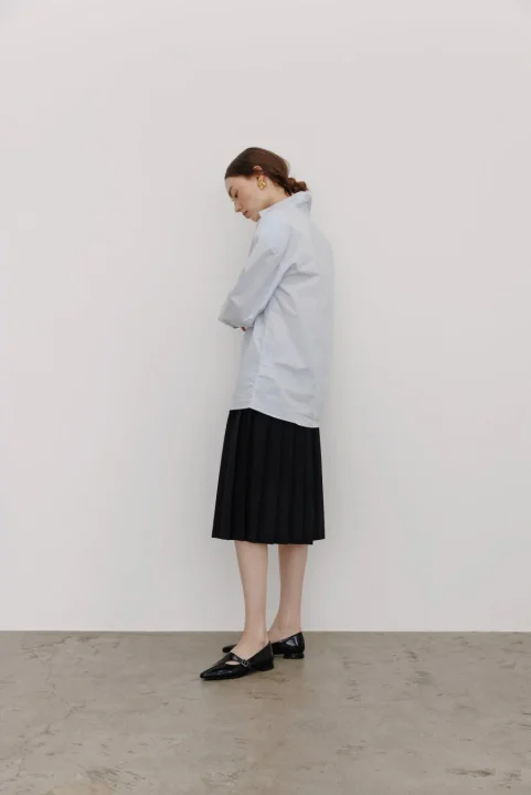 水色のシャツと黒いプリーツスカートを着用した女性の全身像