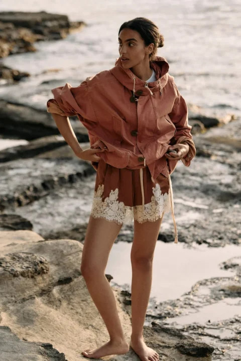 岩場の水辺に立つ女性が写っています。彼女は茶系のカジュアルなジャケットと、裾に白いレースがあしらわれたショートパンツを着用しており、自然体な雰囲気です。