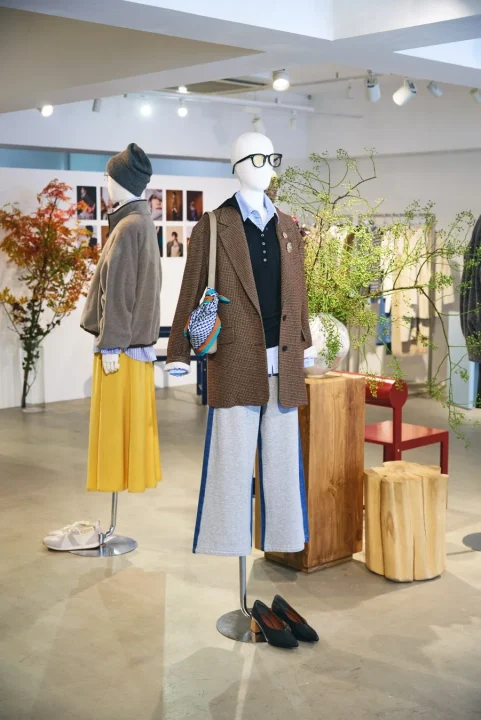 複数のマネキンが秋らしいカジュアルで上品なファッションを展示しているアパレル店舗のディスプレイ。ブラウンのジャケットやイエローのスカートなど、暖色系のアイテムが目を引く。店内には観葉植物や木製スツールが配置され、落ち着いた雰囲気のライフスタイルを提案している。