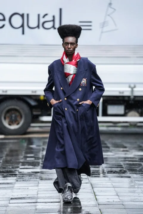 個性的なヘアスタイルのモデルがネイビーのロングコートとスカーフを着用し、雨上がりのような路面を歩いているファッションスナップです。背景にはトラックが見えます。