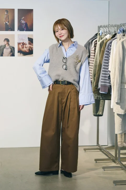 グレーのニットベストと水色のシャツ、茶色のワイドパンツを着用した女性が、笑顔でポーズをとっています。眼鏡をベストにかけ、背後には洋服ラックと写真が飾られています。秋のカジュアルファッションスタイルです。