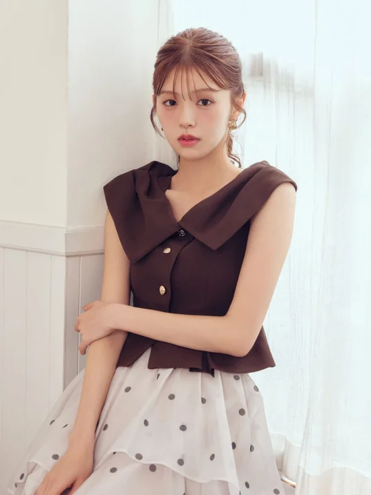茶色の襟付きノースリーブトップスと白いドット柄のティアードスカートを着用した若い女性