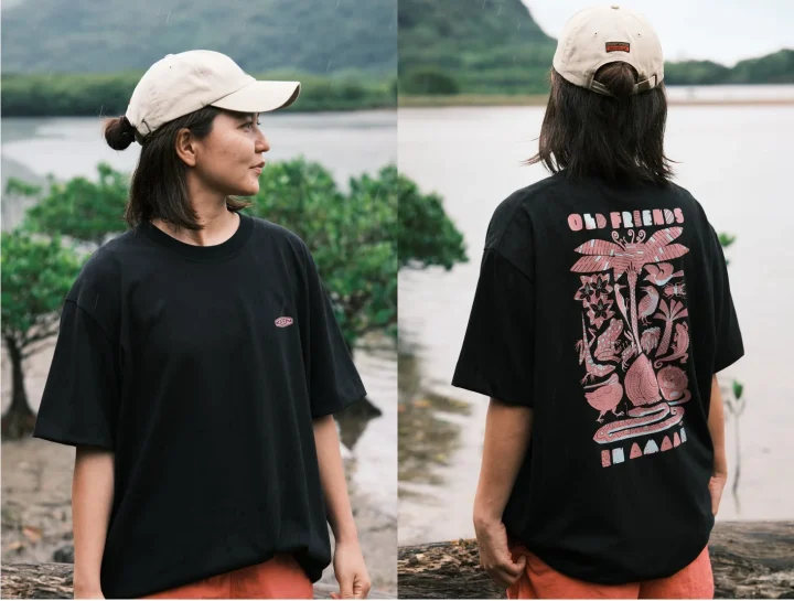 自然豊かな水辺で、黒いTシャツとベージュのキャップを着用した女性がポーズをとっています。Tシャツの背面には「OLD FRIENDS」の文字と動植物をモチーフにした個性的なグラフィックが描かれています。