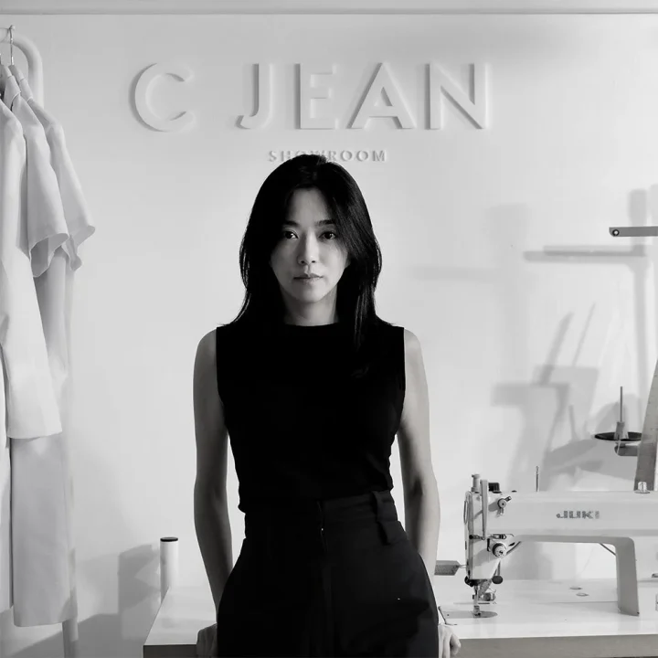 モノクロ写真で、黒い服を着た女性が正面を向いて立っています。背景には「C JEAN SHOWROOM」の文字があり、左にはハンガーにかけられた服、右にはJUKIのミシンが見えます。アパレル関連のショールームでのポートレート写真です。