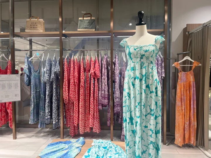 店舗ディスプレイで、マネキンが花柄のリゾートドレスを着用。青、赤、紫など様々な色柄のワンピースが多数並び、ビーチやハワイをイメージさせる夏らしいアパレルが展示されています。