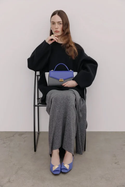 女性が黒いセーターとグレーのスカートを着用し、青とグレーのハンドバッグと青いフラットシューズを合わせている