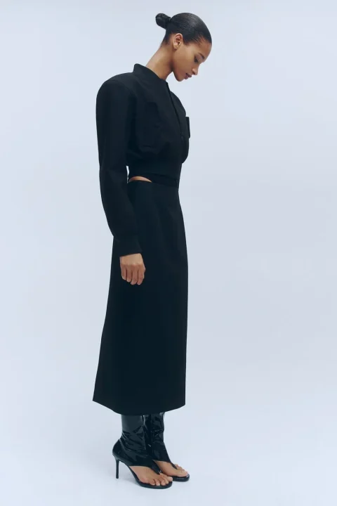 白い背景の前で、黒いクロップド丈ジャケットとミディスカートのセットアップを着用した女性の全身像。特徴的なデザインのサンダルブーツを履き、横向きで下を向いている姿が捉えられています。