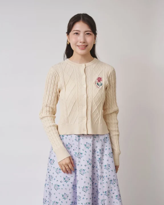 クリーム色のケーブルニットカーディガンと淡い紫の花柄スカートを身につけた若い女性