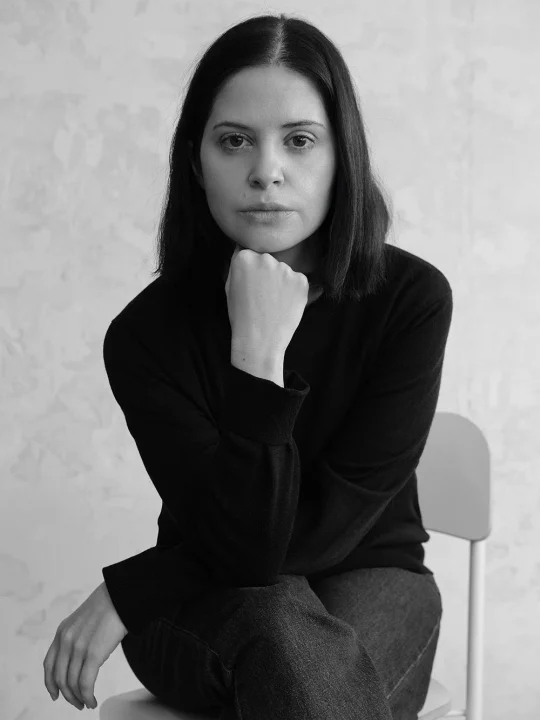 白黒写真に写る女性のポートレート。椅子に座り、顎に手を当てて真剣な表情でカメラを見つめている。黒い服を着用し、シンプルな背景の前で撮影されている。