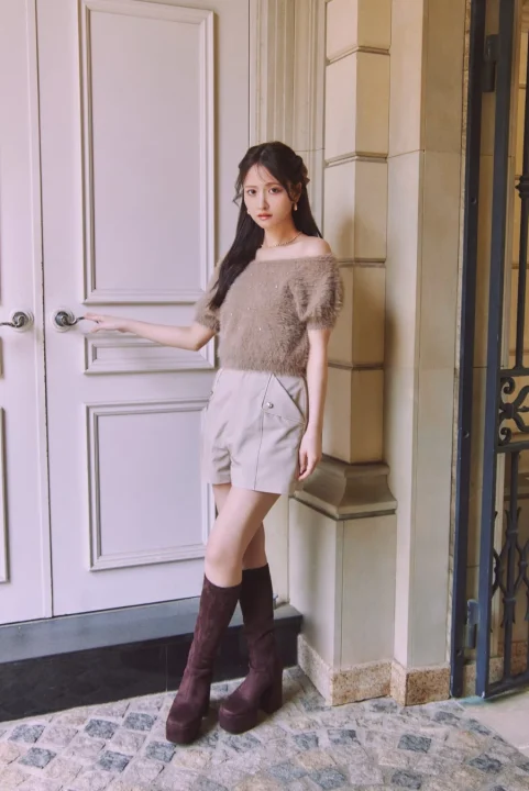 オフショルダーのブラウンニットとベージュのショートパンツ、プラットフォームのロングブーツを着用した女性