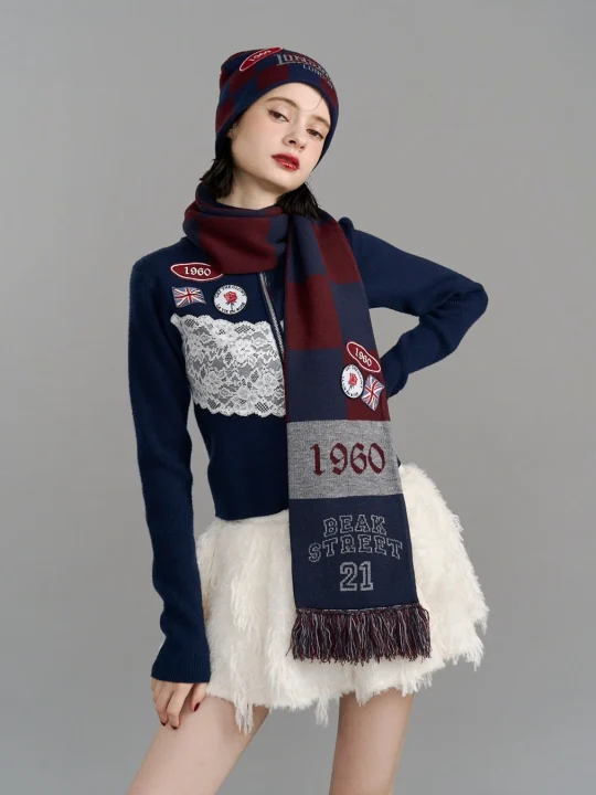 ネイビーのニットウェアと白いふわふわのミニスカートを着用した女性モデルがポーズをとっています。ビーニー帽とマフラーには「1960」や「LONDON」の文字、そしてユニオンジャックなどのワッペンがアクセント