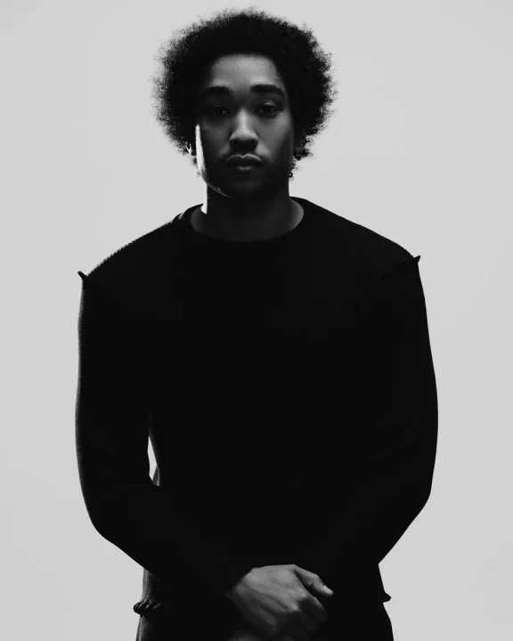 アフロヘアの男性がカメラをまっすぐ見つめる白黒のポートレート。ドラマチックなライティングで顔の一部が強調され、身体には深い影が落ちている。真剣な表情が印象的。