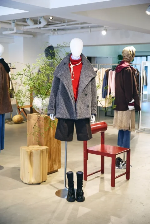 ファッション店舗のディスプレイ。中央のマネキンがグレーのショートコート、赤いトップス、黒のショートパンツ、ブーツを着用。他のマネキンや洋服ラック、植物や木製家具も見える。