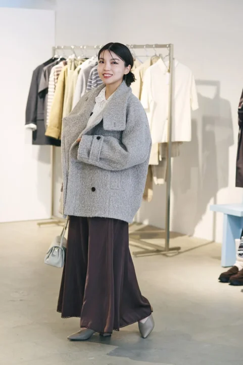 グレーのオーバーサイズコートとブラウンのワイドパンツを着用した女性が、店内で笑顔を見せています。秋冬らしい暖かみのある上品なカジュアルコーディネートです。