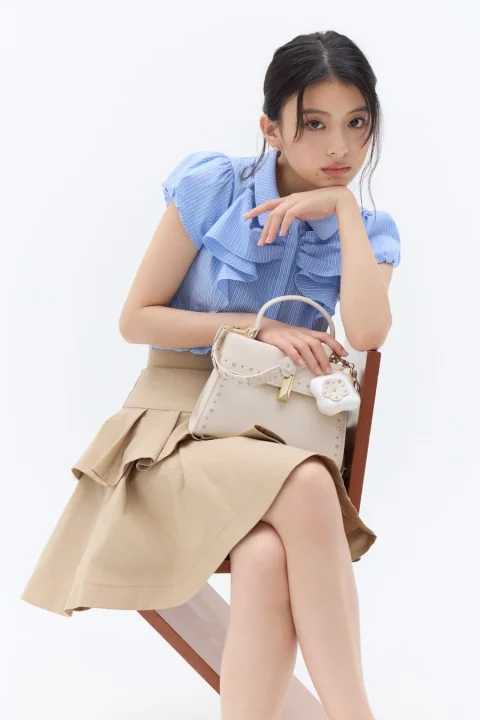 白い背景のスタジオで、椅子に座った若い女性がカメラを見つめています。青いストライプのフリルブラウスとベージュのティアードスカートを着用し、白いハンドバッグを持っています