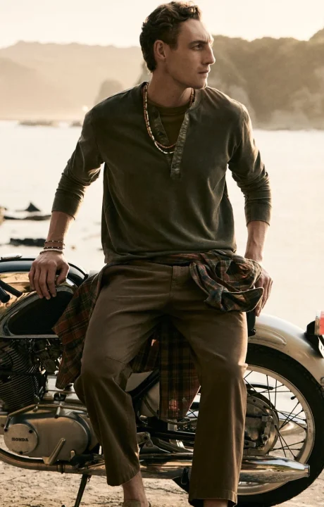 ヴィンテージのオートバイに腰掛けて、遠くを見つめる男性のポートレート。カーキ色のトップスとパンツを着用し、首にはビーズのネックレス、手首にはブレスレットをつけ、腰にはチェック柄のシャツが巻かれています。背景には穏やかな海と山が見え、夕暮れ時の落ち着いた雰囲気が漂っています。