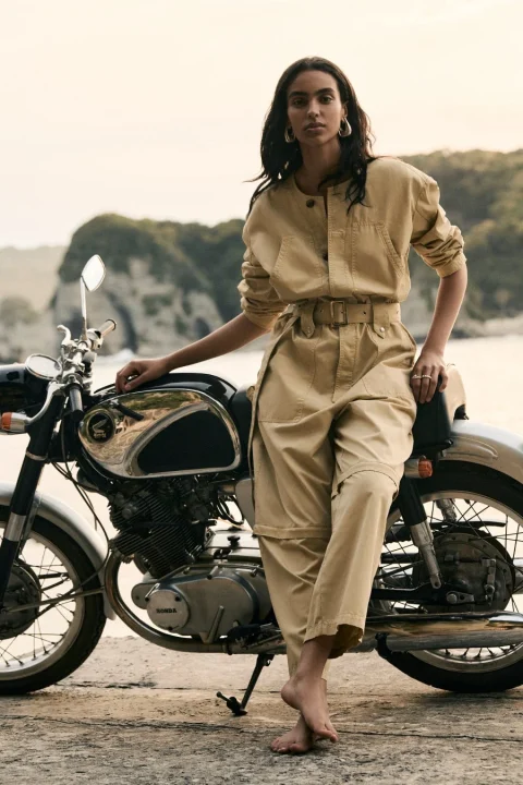 ベージュのジャンプスーツを着た女性が、ヴィンテージのホンダ製オートバイの横に素足で立っています。背景には海と緑豊かな崖が見え、自然の中でのファッションポートレートのような雰囲気です。