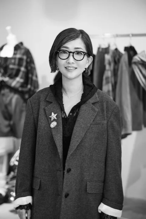 モノクロで撮影された、眼鏡をかけた笑顔の女性のポートレートです。ストライプのブレザーを着用し、左のラペルには星と花のブローチをつけています。背景にはアパレルと思われる衣類が並んでいます。