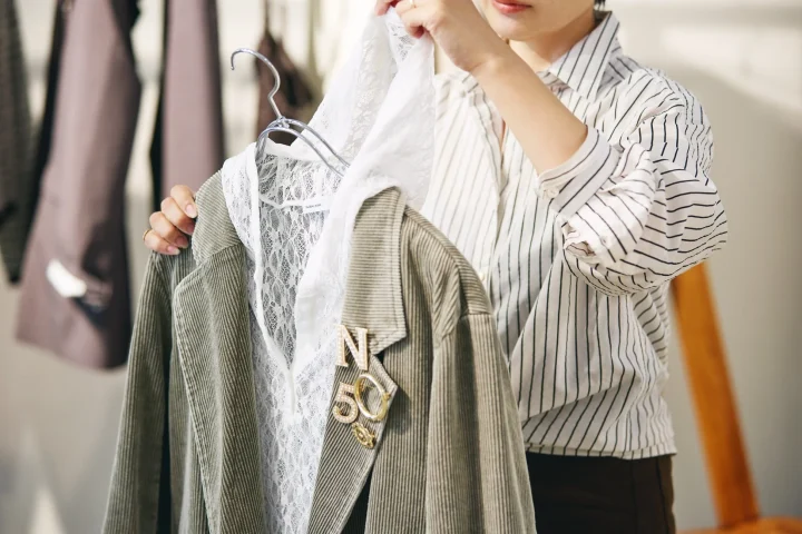 女性がハンガーにかけたコーデュロイジャケットとレーストップスを手に持ち、コーディネートを考えている様子。ジャケットには「N」と「50」のブローチが飾られています。