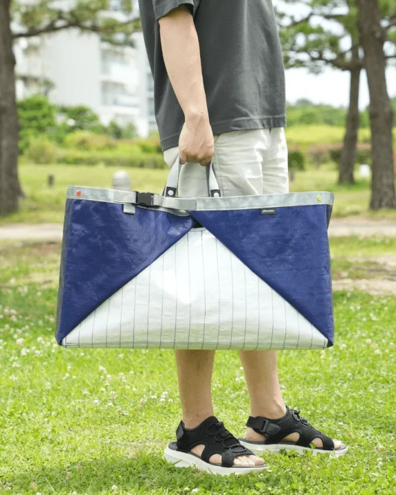 男性が青と白の大きなトートバッグを手に持ち、緑豊かな芝生の上に立っている
