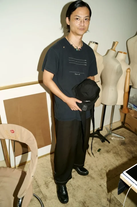 暗いTシャツとワイドパンツ、丸眼鏡をかけたアジア系の男性が、スタジオのような場所で黒い帽子を手に立っています。背景には複数のマネキンや額縁が見えます。