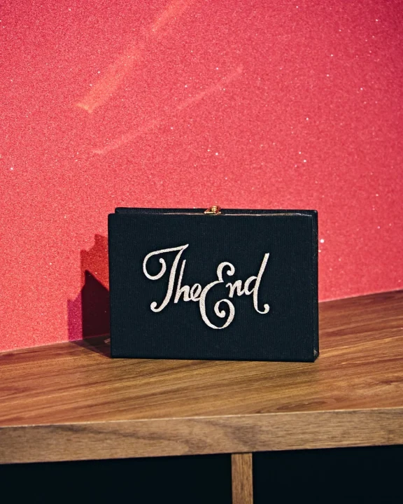 「The End」と書かれた黒いボックス型のクラッチバッグが、光沢のある赤い壁を背景に木製の表面に置かれている写真です。