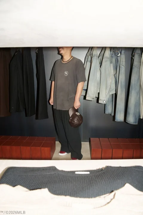 クローゼットのような空間で、ダークカラーのTシャツとワイドパンツを着用した男性が立っています。NYロゴのキャップを手に持ち、背景にはデニムなどの衣類が並んでいます。足元にはチェック柄のスリッポンが見えます。
