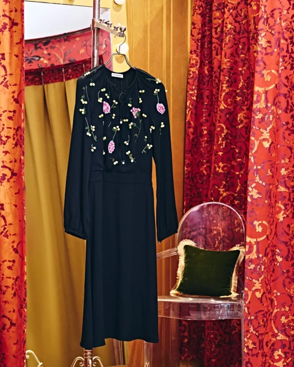 黒い長袖ドレスがハンガーにかかっており、ピンクの花と黄色のドットの刺繍が施されている。背景には豪華な赤い柄のカーテンと金色の壁、透明な椅子と緑のクッションが見える。エレガントなブティックのような雰囲気の画像です。