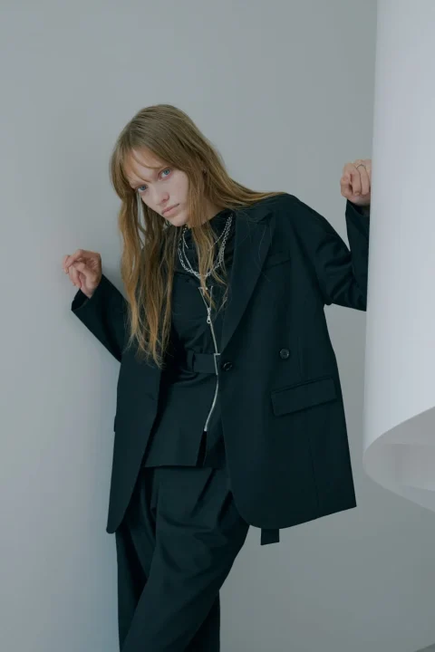 黒いオーバーサイズのスーツを着た女性が、壁に寄りかかってポーズをとっています。
