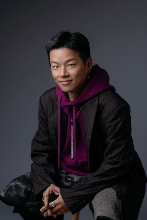 紫のフーディーとダークなブレザーを着たアジア系の男性が、カメラに微笑みかけているポートレート