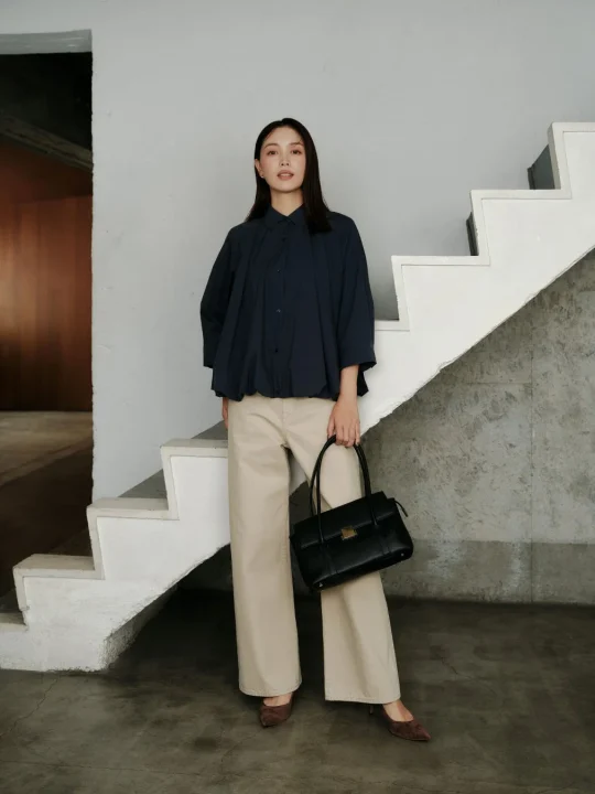 ネイビーのゆったりとしたシャツとベージュのワイドパンツを着用し、黒のハンドバッグを持った女性