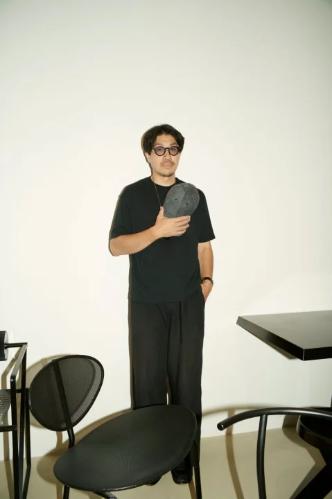 白い背景のスタジオで、黒いTシャツとパンツを着用し、眼鏡をかけた男性が立っています。片手には黒いキャップを持ち、カメラを見つめています。周囲には黒い椅子やテーブルが配置されており、全体的にシンプルで落ち着いた雰囲気のファッションポートレートです。