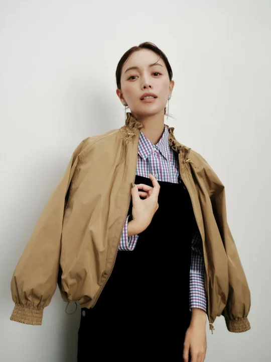 ベージュのボンバージャケットを肩にかけ、チェックシャツと黒いトップスを着用している女性